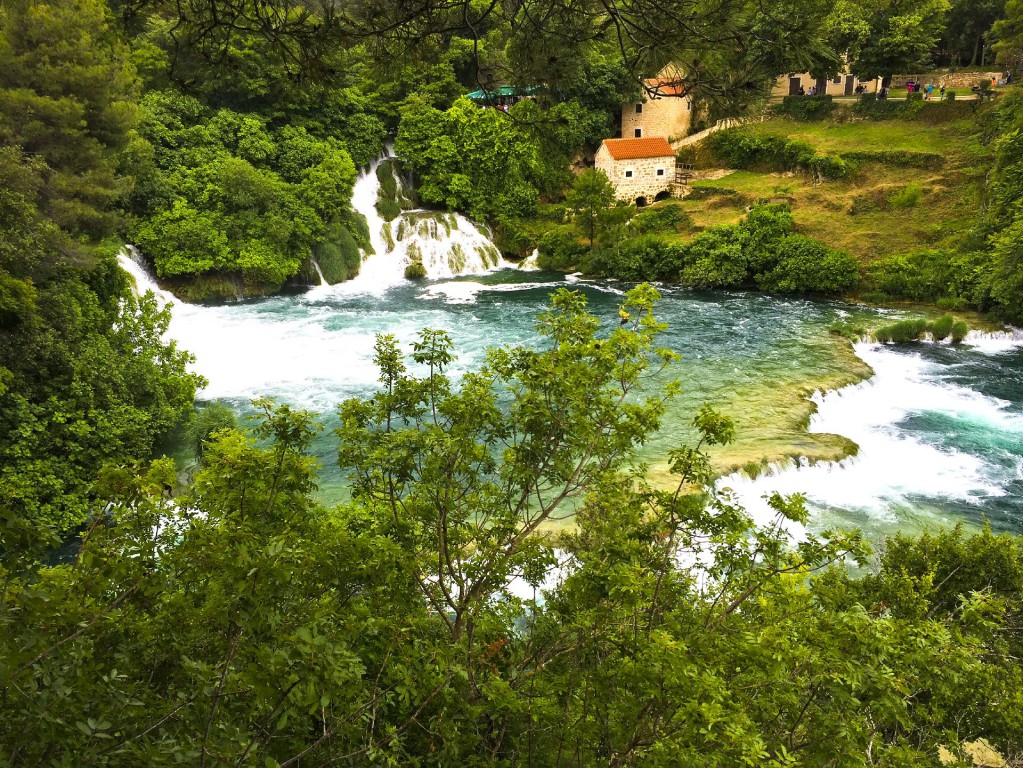 Ein breiter, türkisfarbener Fluss fließt durch ein dicht bewaldetes Tal mit mehreren Wasserfällen im Hintergrund.