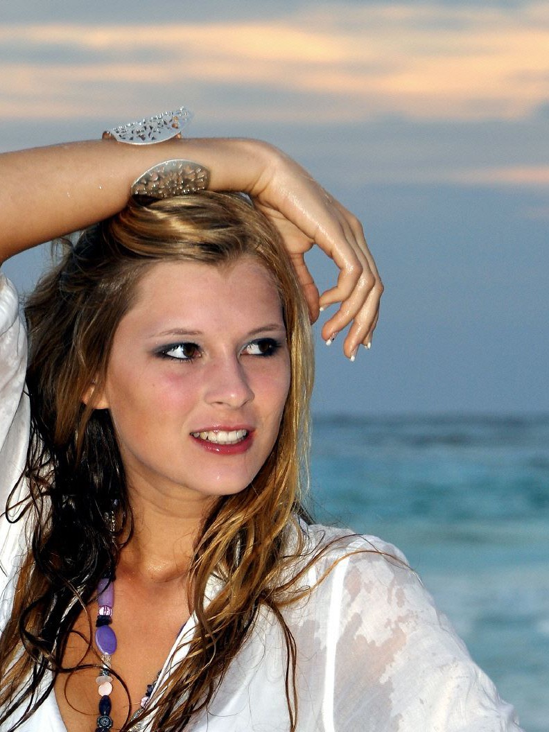 Junge Frau mit langen Haaren steht am Strand und blickt über das ruhige Meer, während sie sich mit einer Hand das Haar berührt.