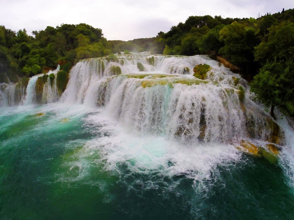 Ein breiter, mehrstufiger Wasserfall stürzt in ein tiefgrünblaues Gewässer, umgeben von dichtem, üppigem Wald.